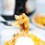 Close-up of a fork holding a bite with melted cheese. In the background, a bowl contains similar ingredients topped with sour cream, reminiscent of a warm chicken lasagna.