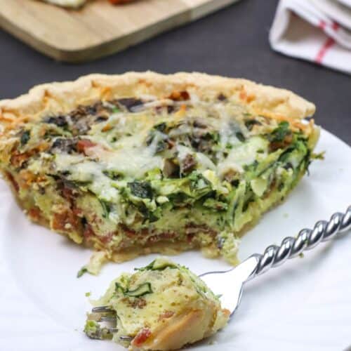 A slice of Spinach and Bacon Quiche with melted cheese on top sits on a white plate, with a fork holding a bite-sized piece in the foreground. Bread and butter are visible in the background.