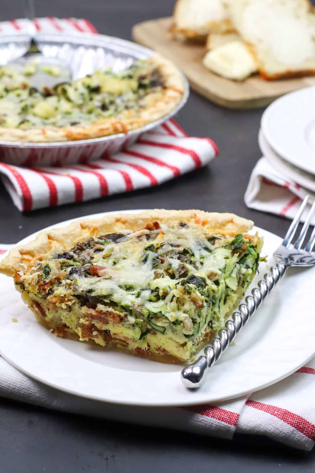 A slice of Spinach and Bacon Quiche with melted cheese sits on a white plate, accompanied by a fork, with more quiche and sliced bread in the background.