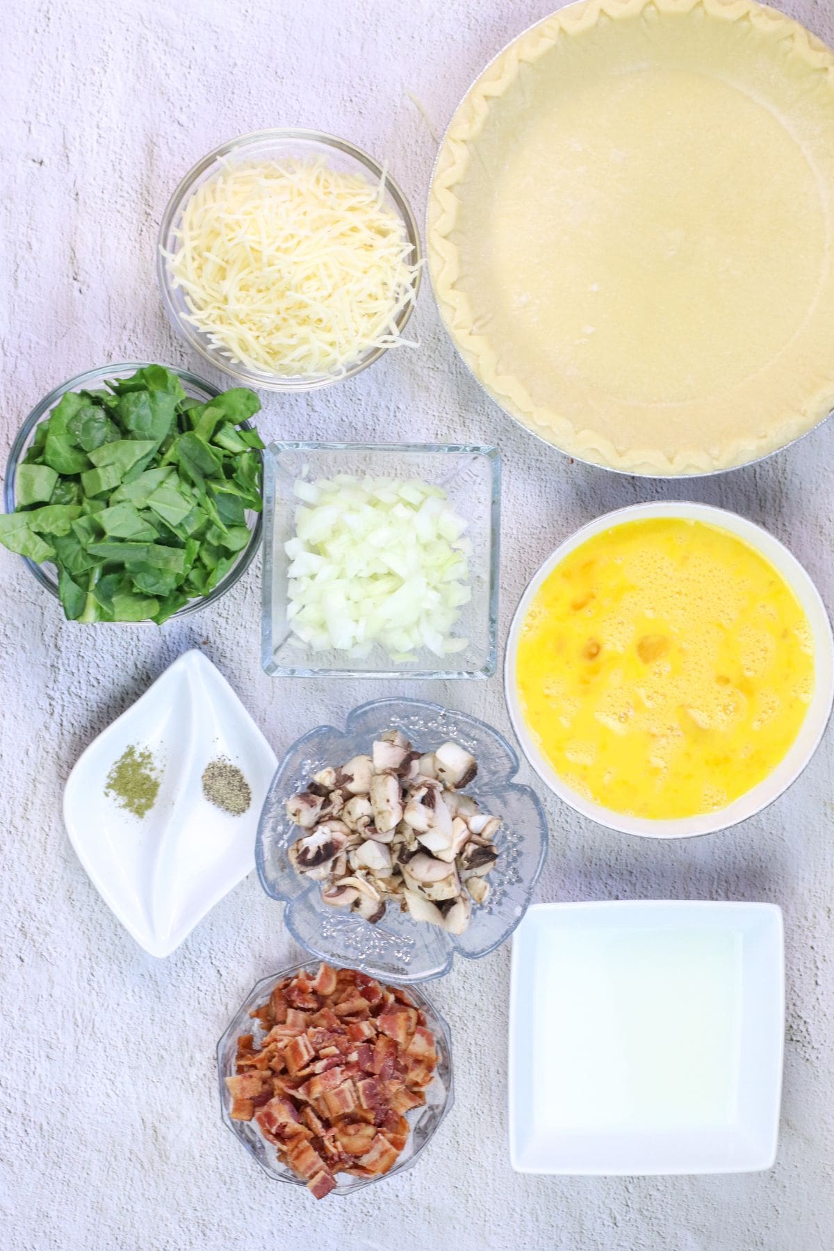 Ingredients for a Spinach and Bacon Quiche arranged on a light surface, including shredded cheese, pie crust, spinach, chopped onion, beaten eggs, spices, mushrooms, cooked bacon, and milk.