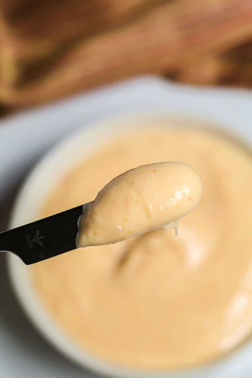 A close-up of a knife spreading creamy, light orange Sriracha Aioli over a bowl filled with the same spicy sauce.