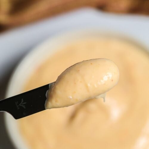 A close-up of a knife spreading creamy, light orange Sriracha Aioli over a bowl filled with the same spicy sauce.