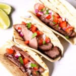 Three grilled Steak Street Tacos with diced tomatoes, onions, and cilantro in soft tortillas, placed on a white surface with lime wedges nearby.