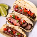 Three delicious Steak Street Tacos with grilled steak, chopped tomatoes, onions, and cilantro on flour tortillas, served alongside fresh lime wedges.