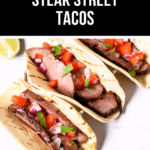Three mouthwatering steak street tacos, perfectly grilled with diced tomatoes, onions, and cilantro atop soft tortillas, all presented on a pristine white surface.