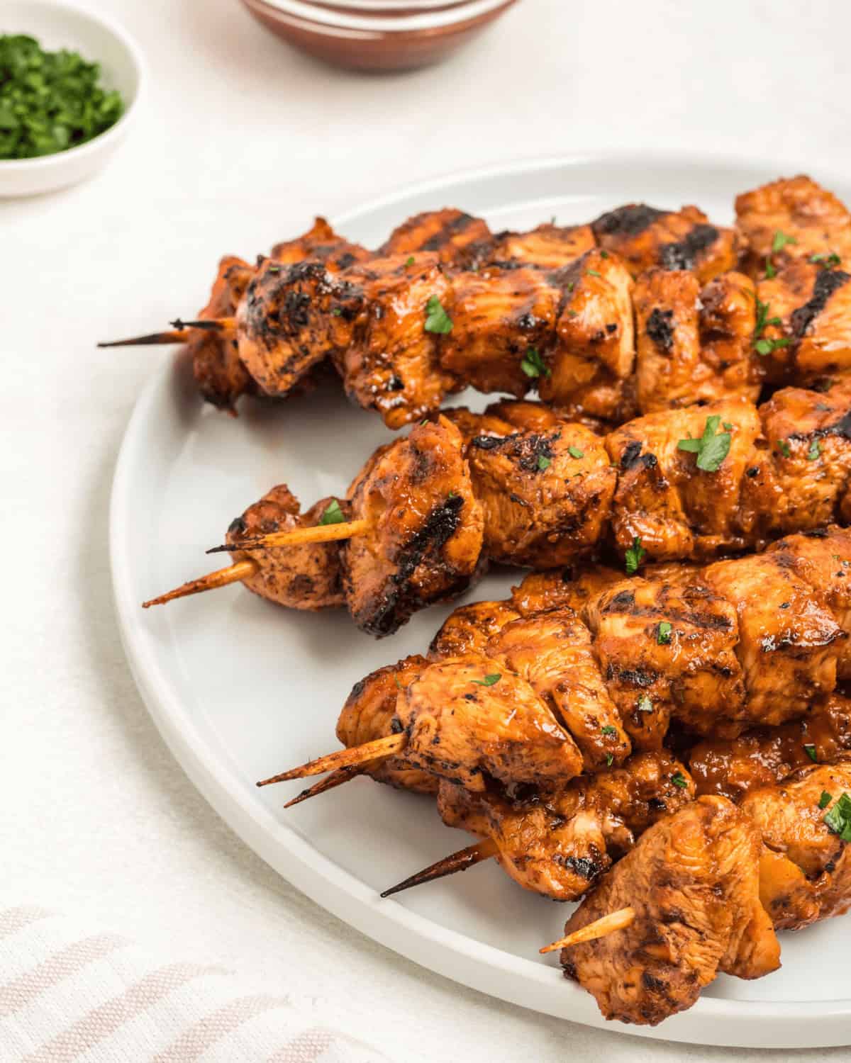 Three skewers rest on a white plate, garnished with chopped herbs.