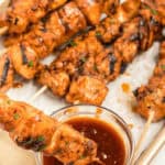 Grilled chicken on a stick arranged on a parchment-lined tray with a small bowl of dipping sauce in the foreground.
