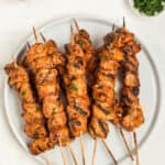 A plate with six chicken on a stick skewers garnished with herbs, accompanied by a small dish of red sauce and a small bowl of chopped green herbs.