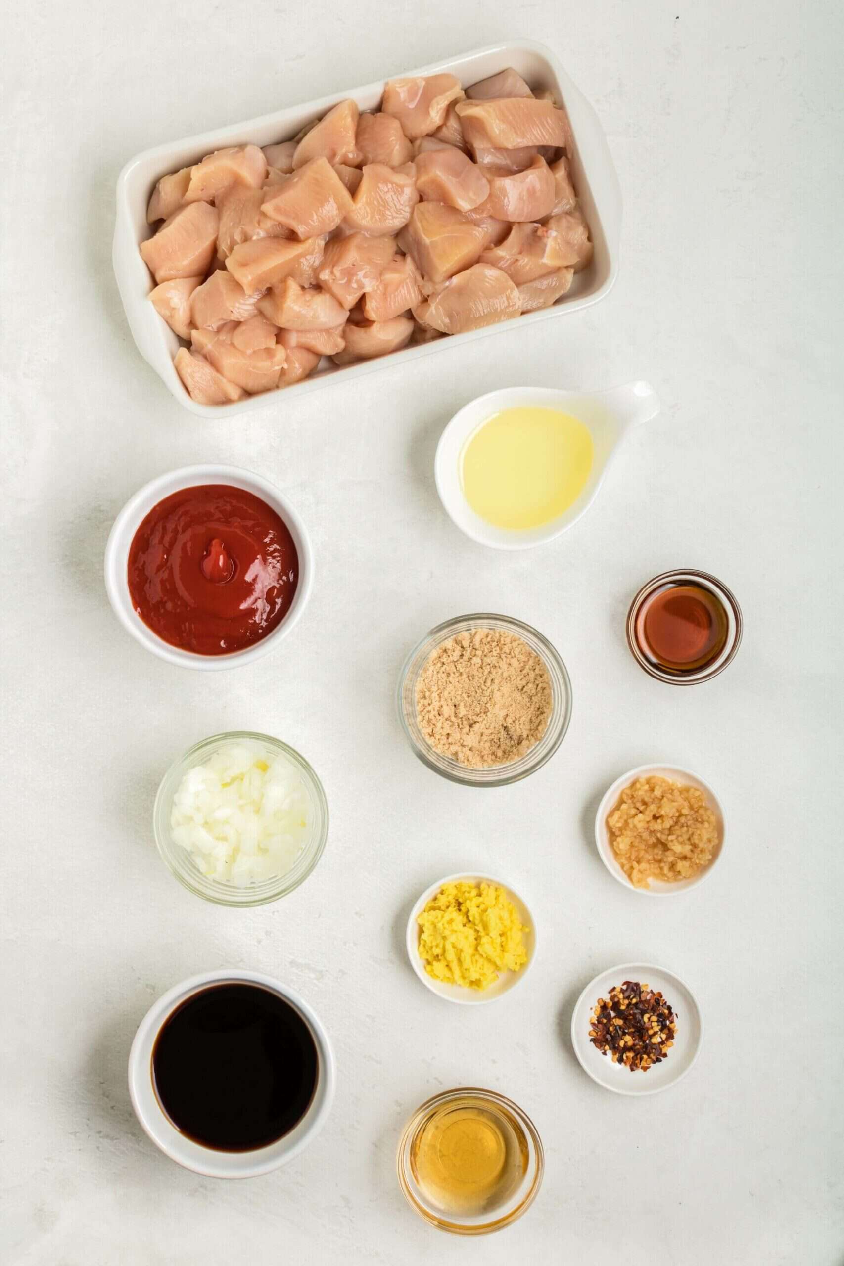 Ingredients for a delicious chicken on a stick recipe arranged on a white surface, including diced chicken, ketchup, garlic, soy sauce, honey, crushed red pepper, minced ginger, eggs, lemon juice, breadcrumbs, and vinegar.