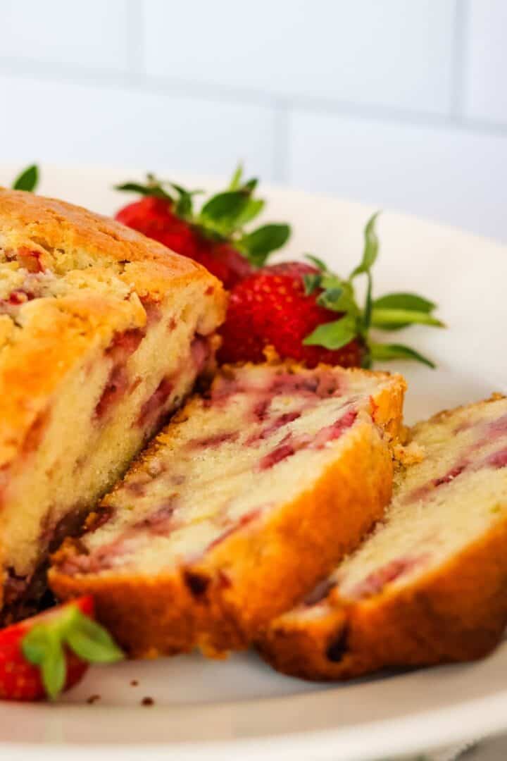 A close-up of sliced strawberry bread from our favorite Strawberry Bread Recipe, with visible strawberry pieces, served on a white plate and garnished with whole strawberries.