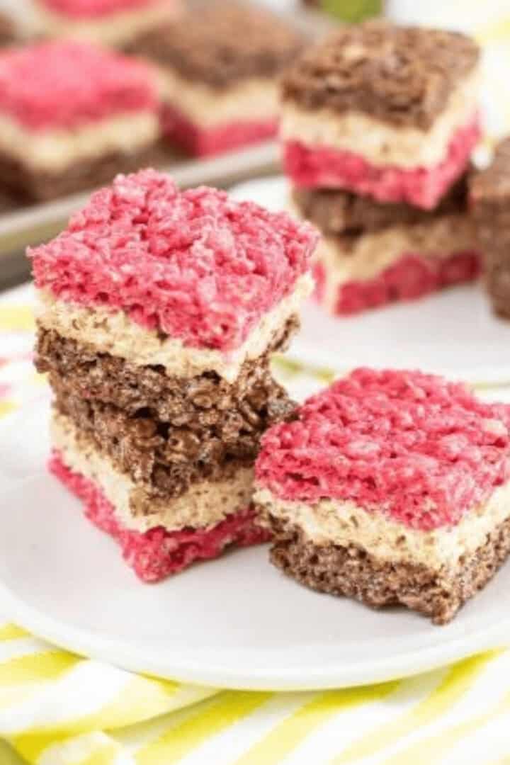 Stacked Chocolate Rice Krispie Treats in pink, white, and brown layers rest invitingly on a white plate. More delicious treats are visible in the background, promising a burst of flavor with every bite.