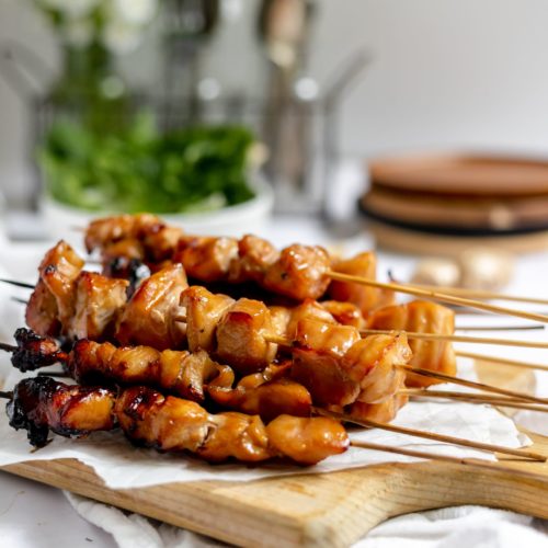On a wooden cutting boards there are a stack of Teriyaki Chicken on a Stick.