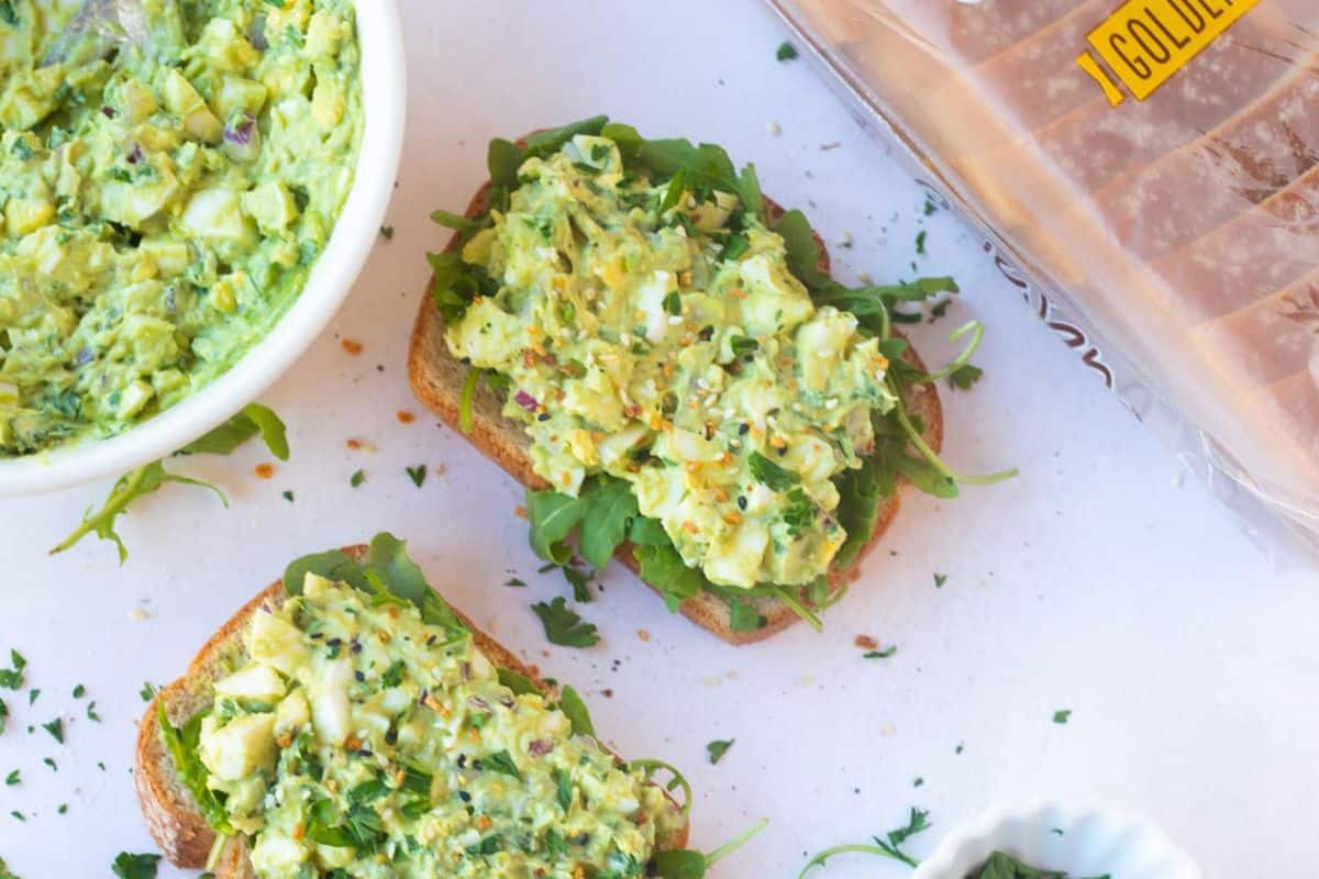 Two slices of toast topped with avocado egg salad and greens are on a white surface, accompanied by a bowl of salad and a loaf of sliced bread, perfect for pairing with easy egg salad recipes.