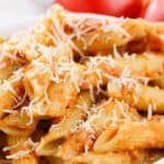 A close-up of penne pasta with vodka cream sauce and grated cheese in a white bowl, with tomatoes in the background.