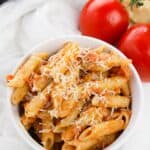 A white bowl filled with penne pasta in rich vodka cream sauce, topped with grated cheese, sits next to two whole tomatoes on a white cloth.