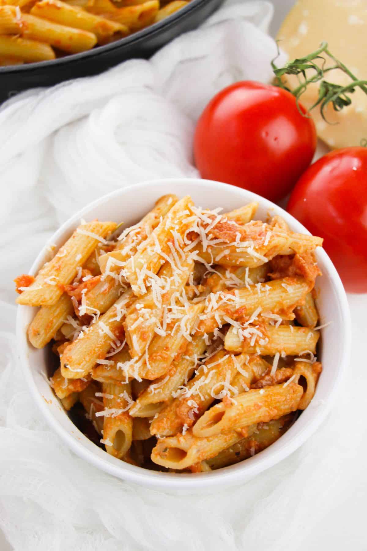 A white bowl filled with penne pasta in rich vodka cream sauce, topped with grated cheese, sits next to two whole tomatoes on a white cloth.