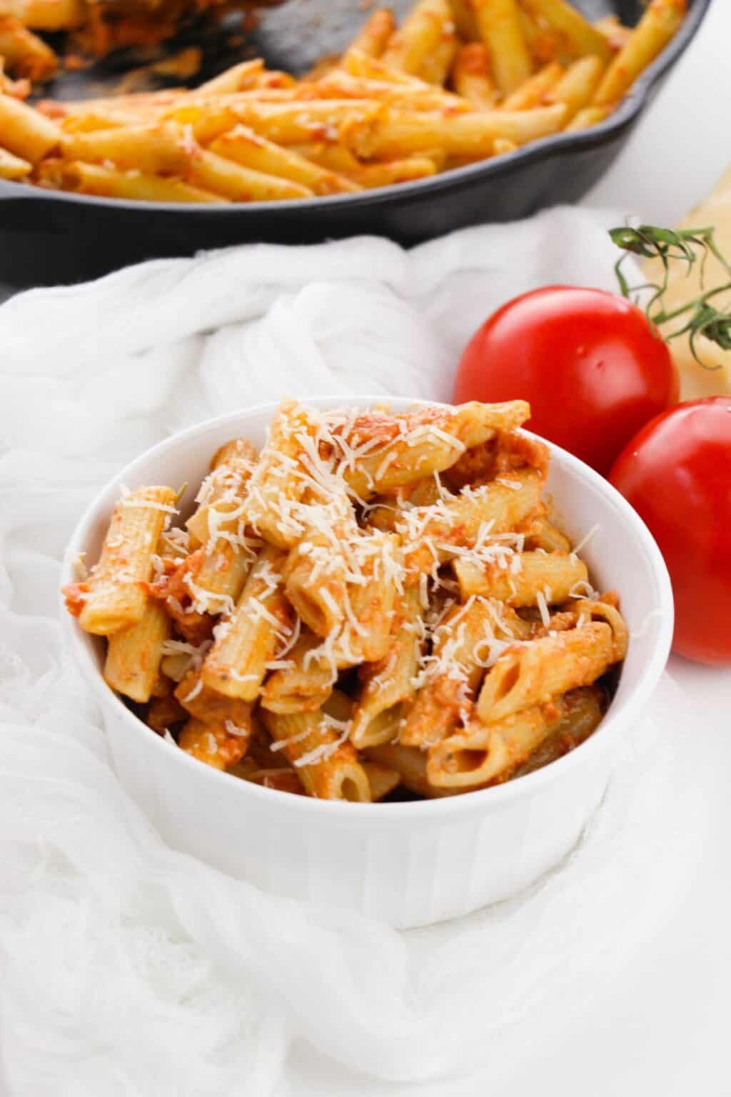A white bowl filled with penne topped with grated cheese, sits next to two whole tomatoes and a skillet of pasta.