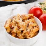 A white bowl filled with penne topped with grated cheese, sits next to two whole tomatoes and a skillet of pasta.
