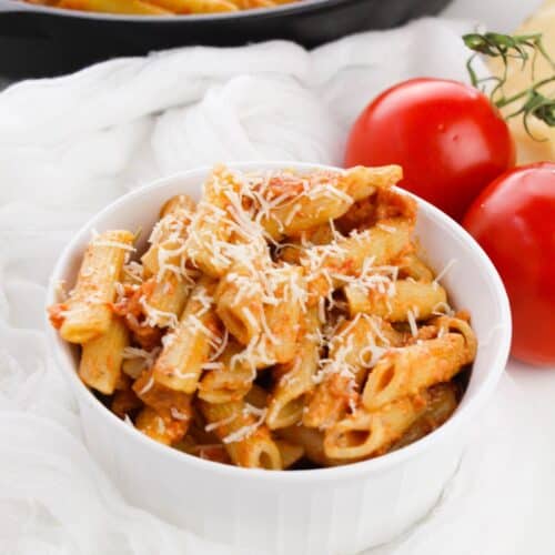 A white bowl filled with penne topped with grated cheese, sits next to two whole tomatoes and a skillet of pasta.