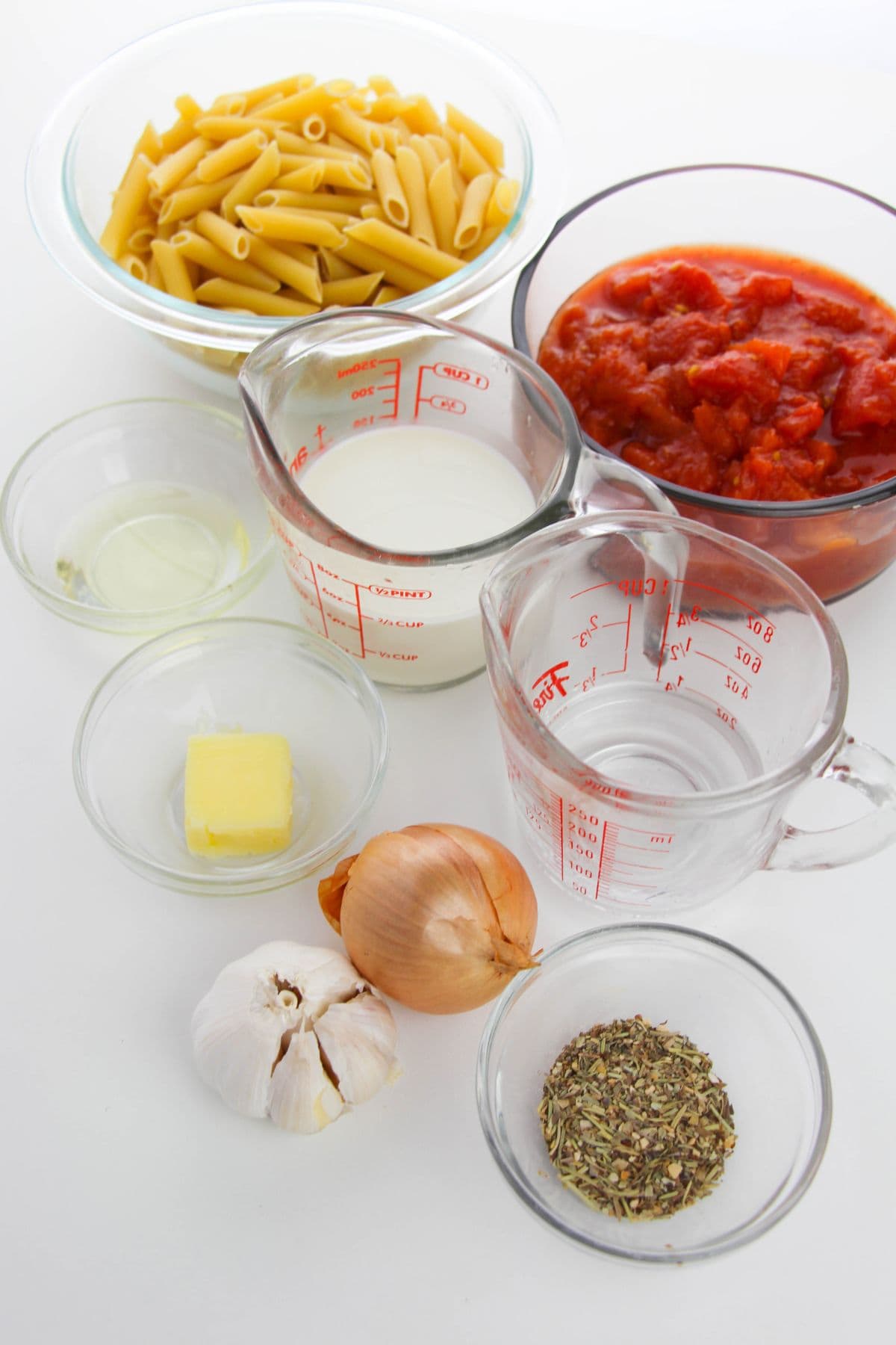 Various ingredients are arranged on a white surface, including dry pasta, tomato sauce, milk, water, oil, butter, an onion, garlic, a bowl of dried herbs—perfect essentials for creating a rich Vodka Cream Sauce.