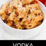 A white bowl filled with penne pasta tossed in rich Vodka Cream Sauce and topped with grated cheese, with fresh tomatoes in the background. Text at bottom reads "Vodka Cream Sauce.