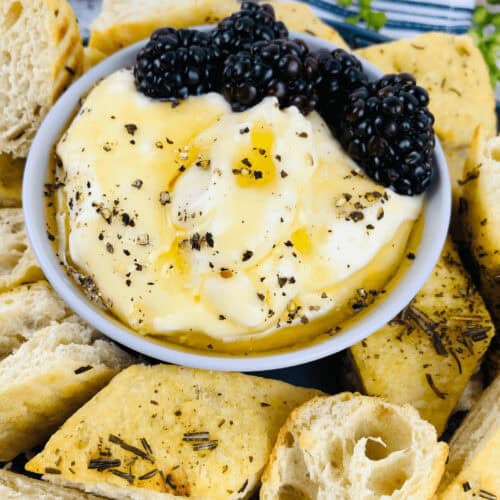A bowl of whipped ricotta dip, creamy and smooth, topped with blackberries and pepper, surrounded by sliced bread pieces garnished with herbs.