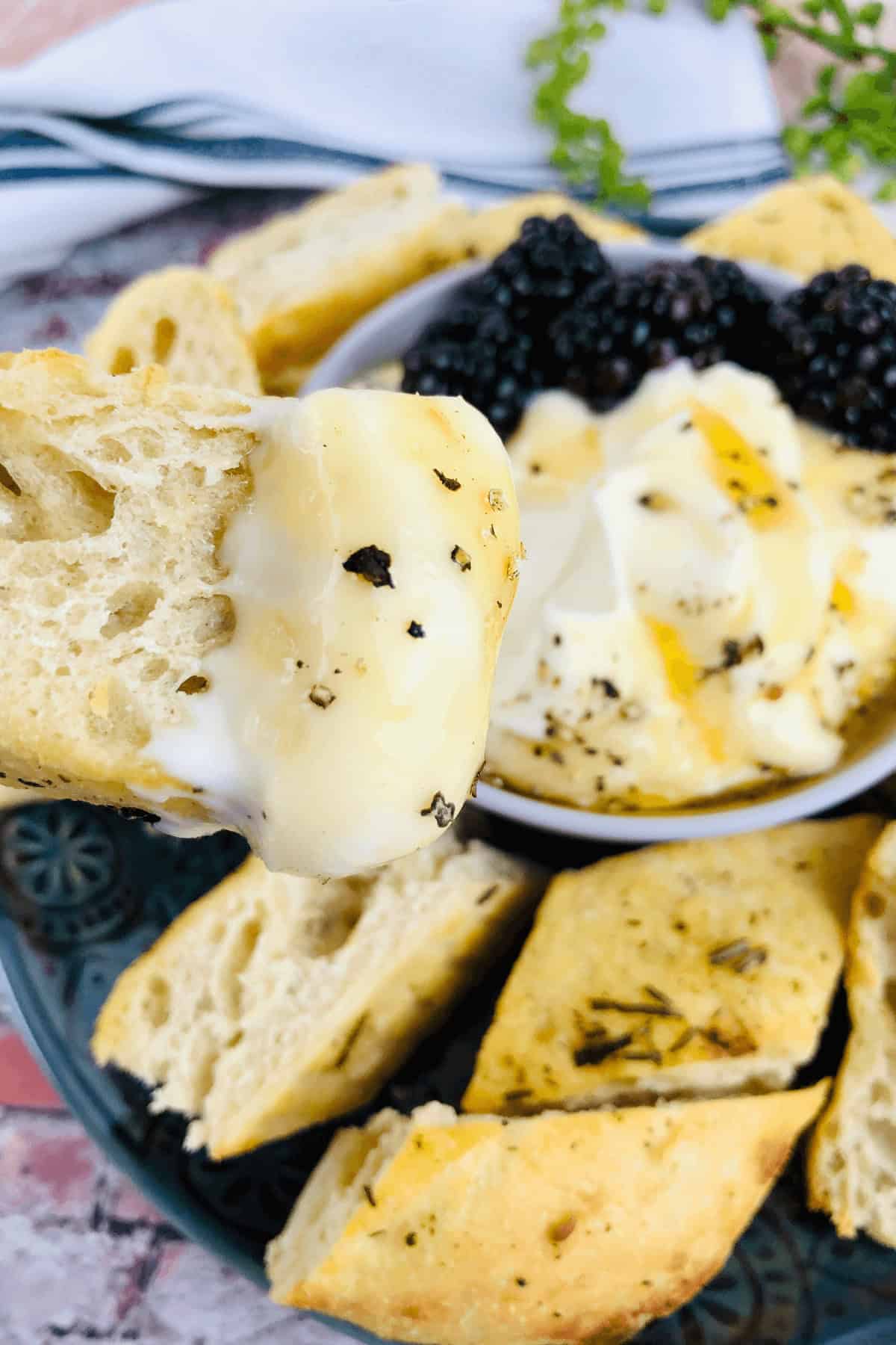 A bowl of whipped ricotta dip, topped with black pepper and juicy blackberries, is surrounded by slices of bread. One slice is being held up close to the camera.
