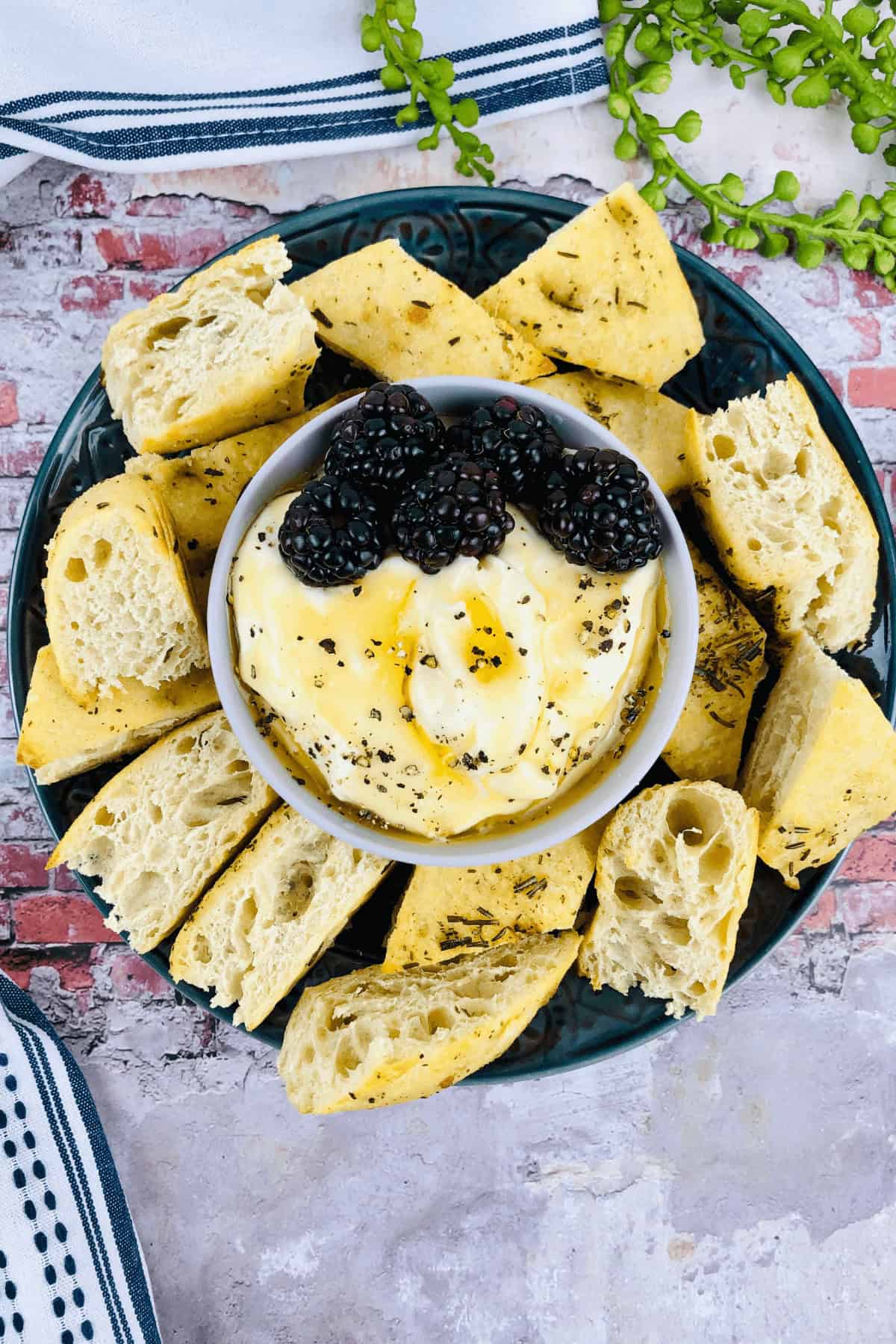 A plate with sliced focaccia encircles a bowl of burrata, blackberries, and honey, garnished with herbs—a delightful companion to a whipped ricotta dip.