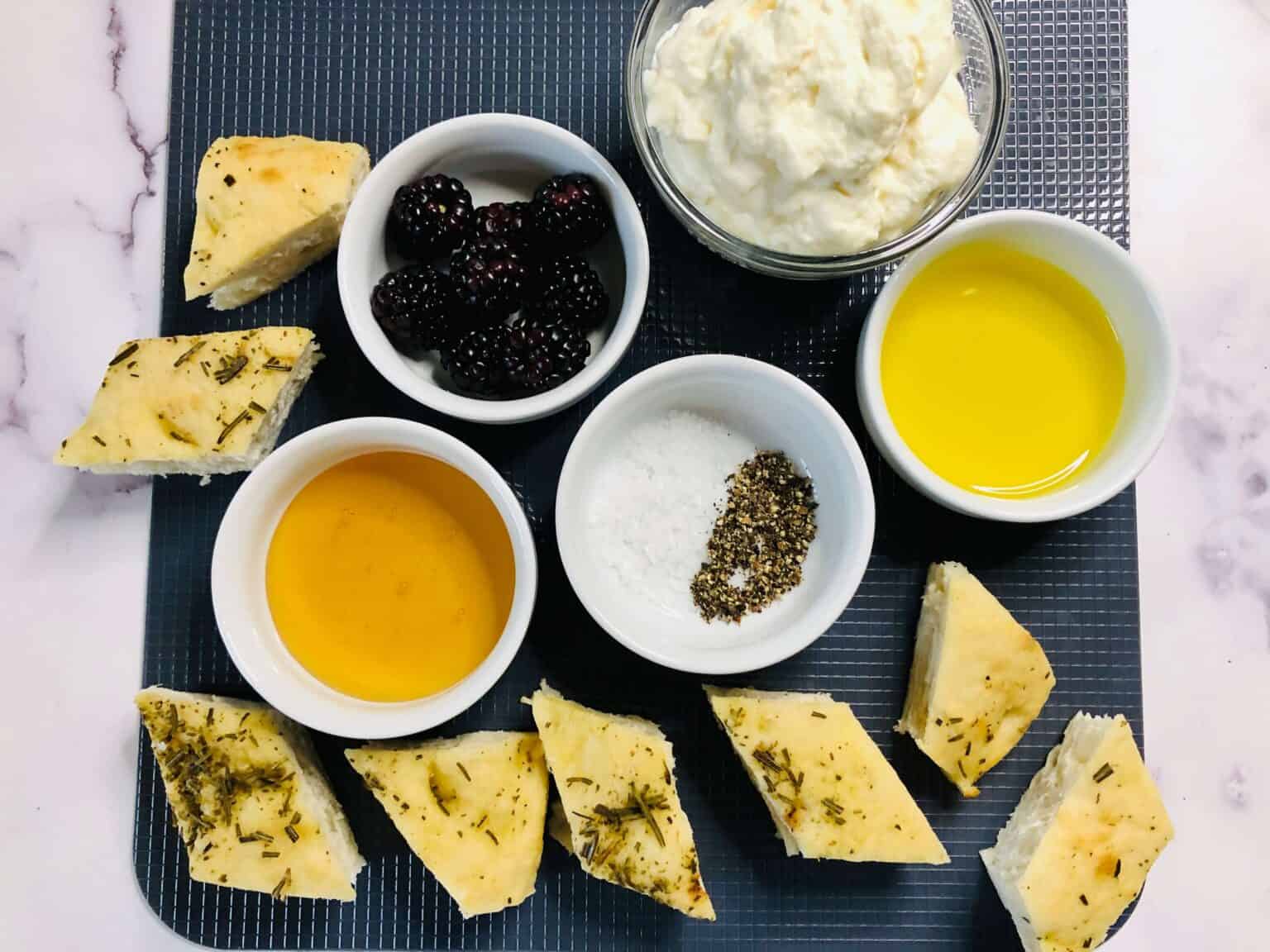 Food items arranged on a dark mat include bread pieces, blackberries, whipped cream, olive oil, and honey surrounding a dish with salt and pepper, perfect for pairing.
