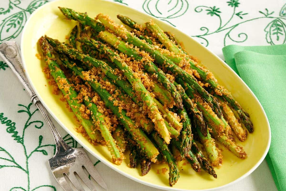 Crispy air-fried asparagus spears topped with golden breadcrumbs, served on a yellow oval dish with a vintage fork on the side.