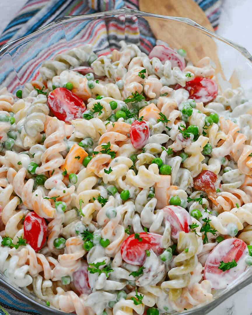 A glass bowl filled with bacon pasta salad featuring rotini, cherry tomatoes, peas, cubed cheese, and parsley, all coated in a creamy ranch dressing. A wooden spoon and striped cloth are in the background.