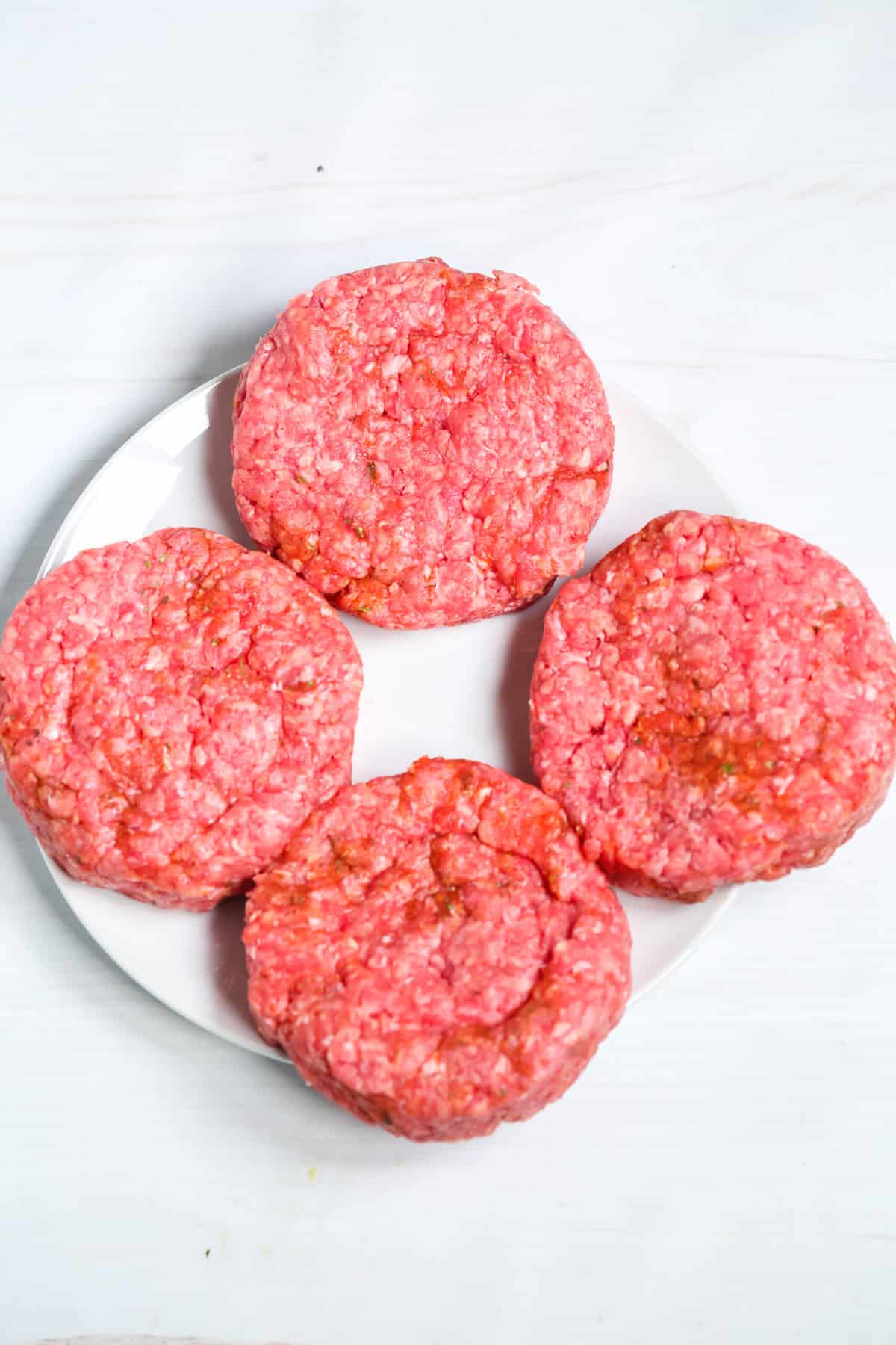 Four raw ground beef patties, soon to become delicious cast iron burgers, are arranged on a white plate against a white background.