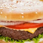 A close-up of a Cast Iron Burger featuring a sesame seed bun, lettuce, a beef patty, melted cheese, tomato slices, and onion rings.