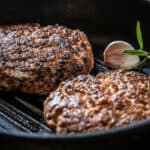 Two Cast Iron Burgers sizzle in a black cast iron skillet beside a clove of garlic and a sprig of rosemary.