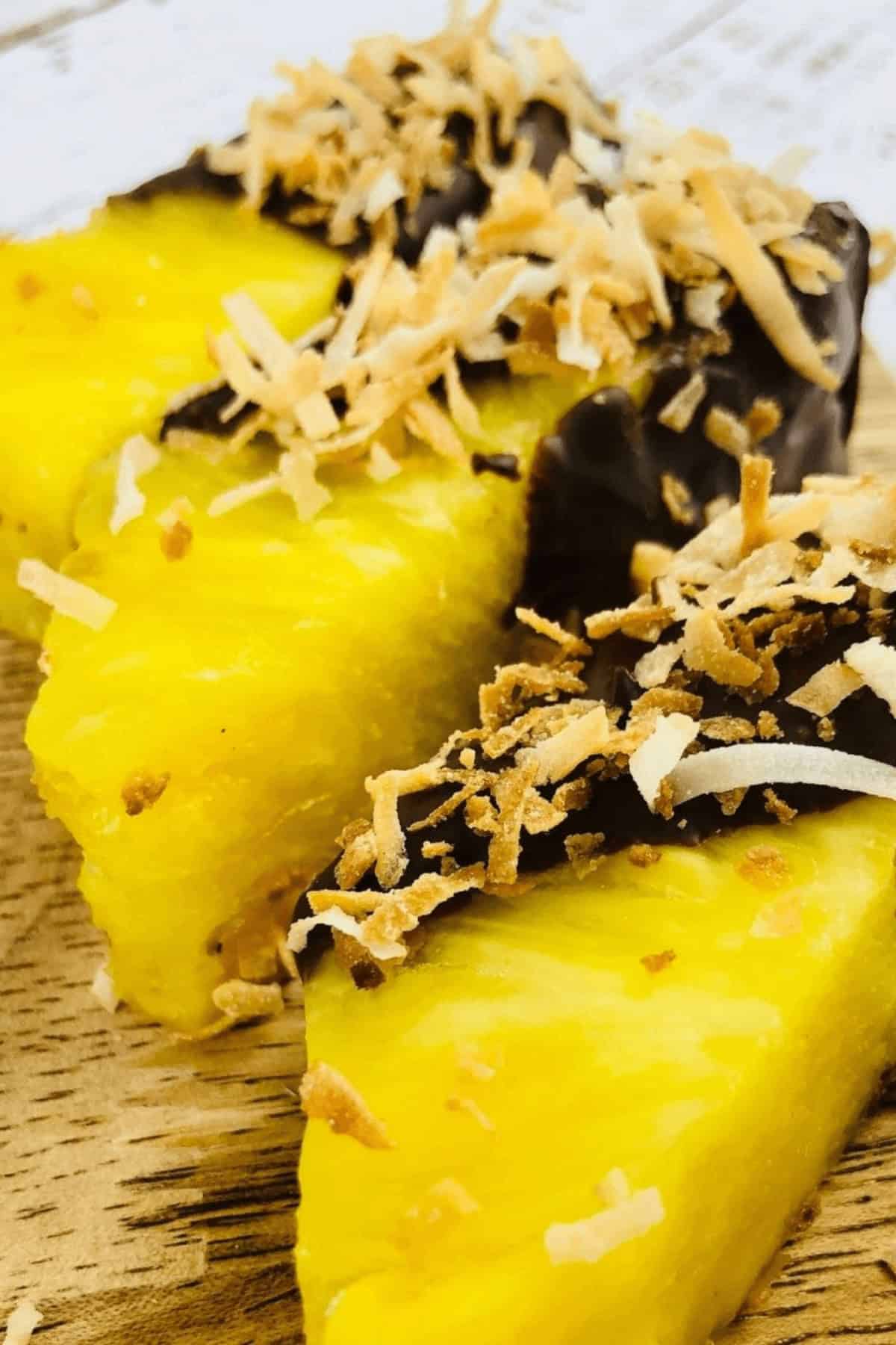 Close-up of chocolate-covered pineapple wedges dipped in melted chocolate and topped with toasted coconut flakes, placed on a wooden surface.