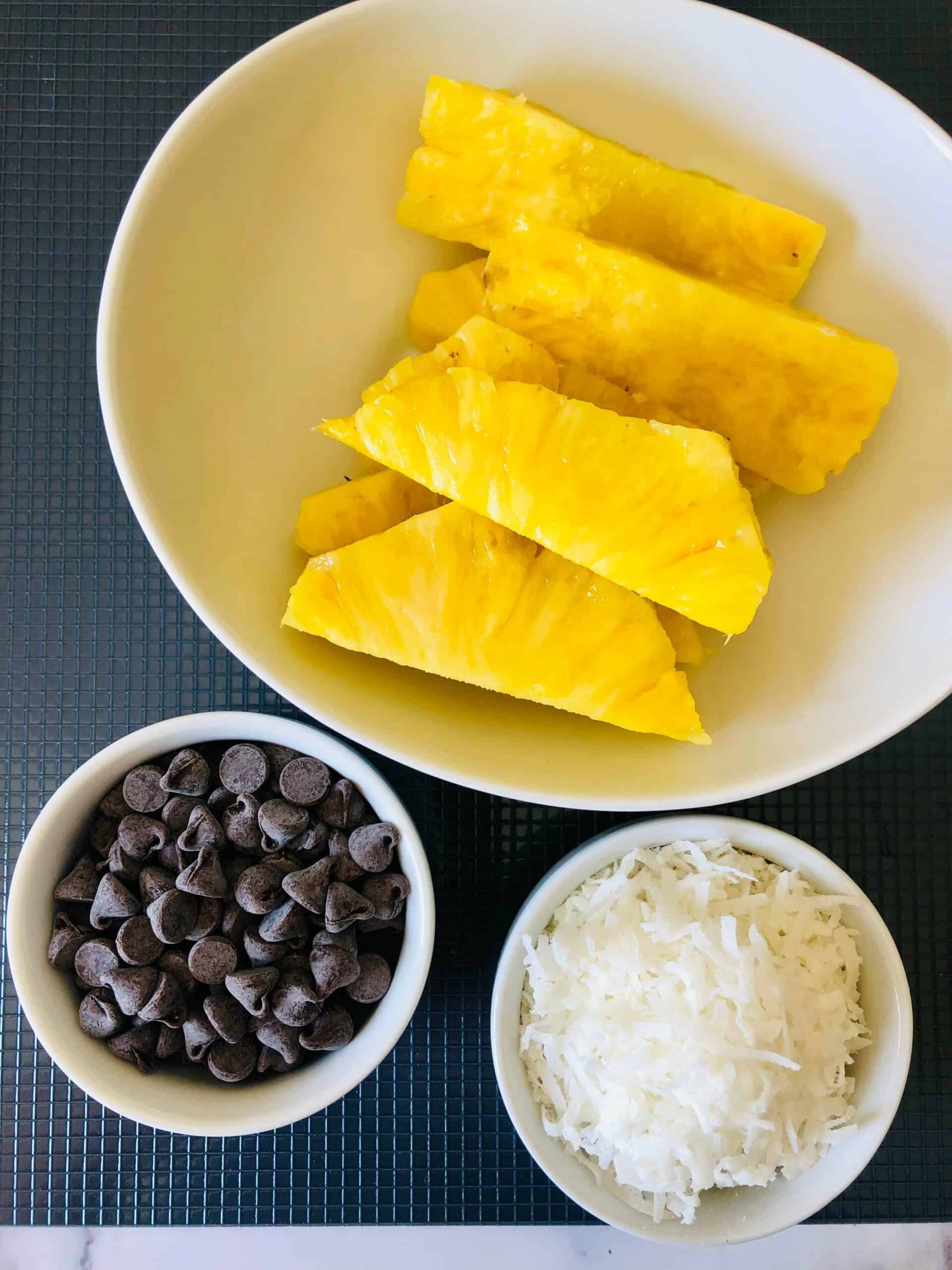 A white bowl with pineapple slices, a small bowl of chocolate chips, and a small bowl of shredded coconut are arranged on a dark surface.