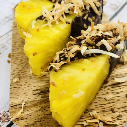 A wooden cutting board with pineapple covered in chocolate and coconut.