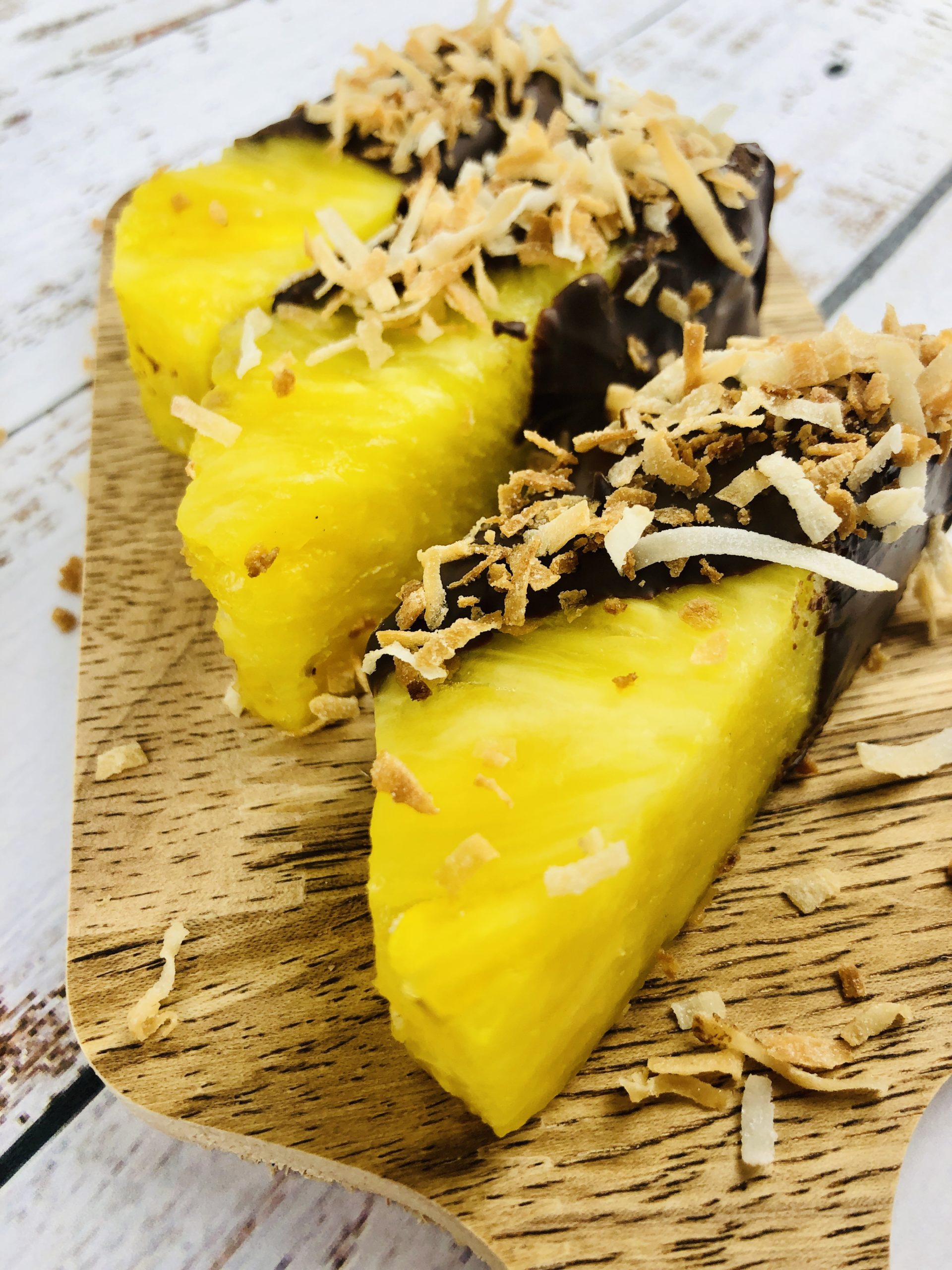 A wooden cutting board with pineapple covered in chocolate and coconut.