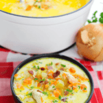 A bowl of creamy Cracked Chicken Soup brimming with vegetables and bacon sits invitingly on a red and white checkered cloth, accompanied by a larger pot of soup and an onion in the background.