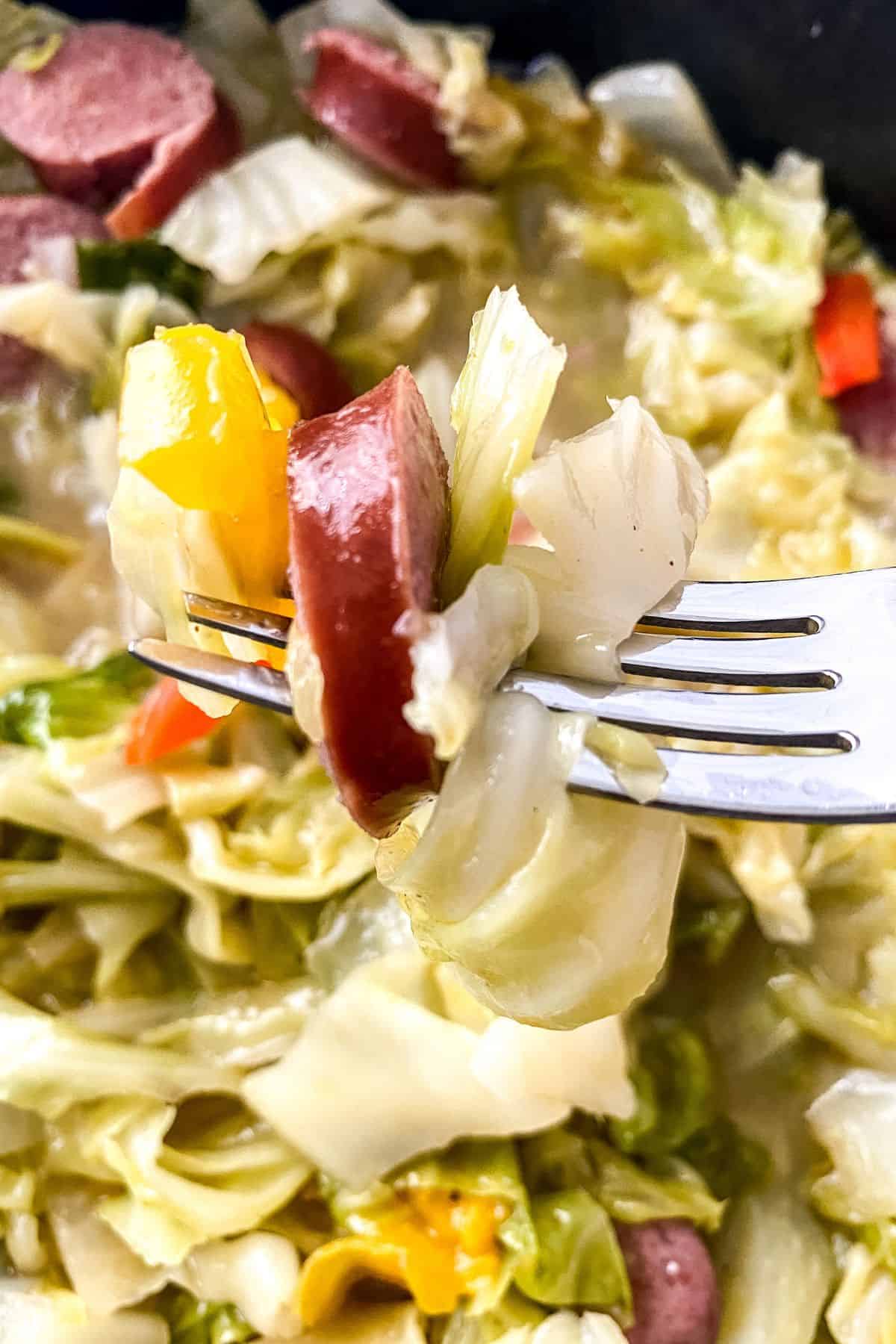 A close-up of a fork holding a bite of Fried Cabbage and Sausage with bell pepper, with more of the tasty dish visible in the background.