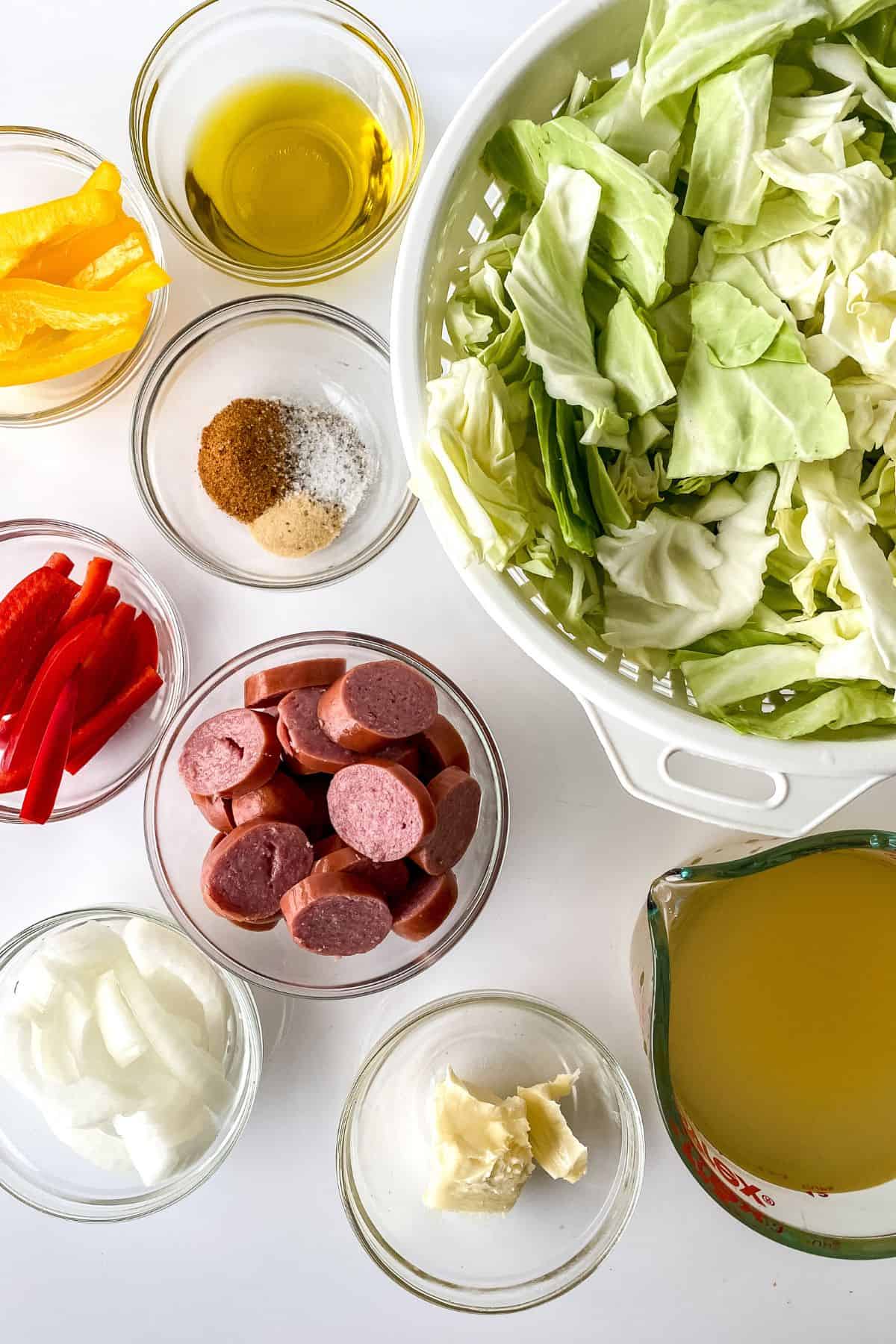 Ingredients for Fried Cabbage and Sausage arranged on a white surface, including chopped cabbage, sliced sausage, bell peppers, onion, garlic, spices, olive oil, and broth.