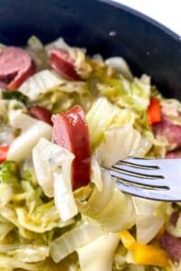 A fork holding a bite of Fried Cabbage and Sausage, with more cabbage, sausage, and vegetables visible in the background.