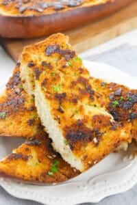 Three slices of homemade garlic bread with a golden, crispy crust are stacked on a white plate, garnished with chopped herbs.