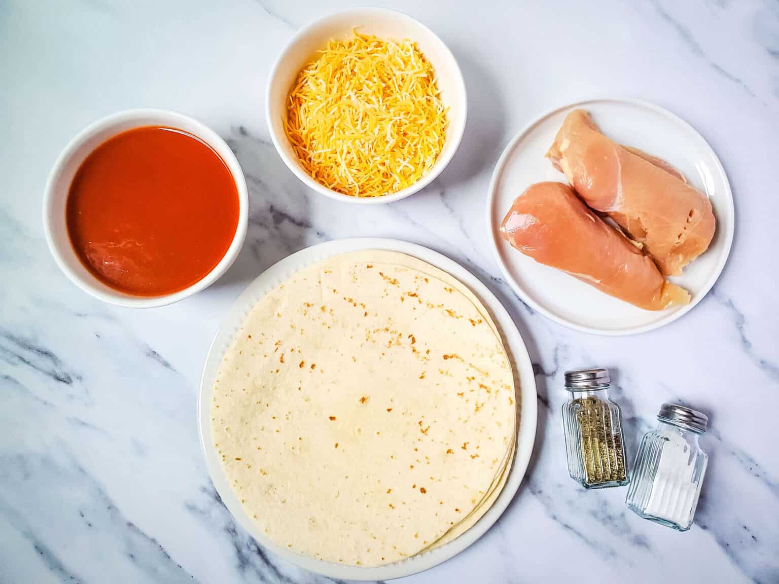 A preparation of lazy enchiladas featuring chicken breasts, shredded cheese, tortillas, enchilada sauce, and salt and pepper shakers arranged on a marble surface.