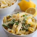 A bowl of Lemon Asparagus Pasta with green beans, peas, and grated cheese, accompanied by a fresh lemon and another bowl of pasta in the background.