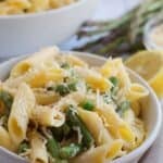 A bowl of ziti with peas, and grated cheese sits on a white surface, accented by a fresh lemon and asparagus stalks in the background.