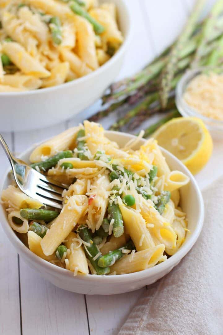 A bowl of Lemon Asparagus Pasta features penne mixed with asparagus and topped with grated cheese, accompanied by a fork, lemon, and fresh asparagus spears in the background.