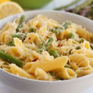 A bowl of Lemon Asparagus Pasta features penne mixed with asparagus and topped with grated cheese, with fresh lemon and asparagus visible in the background.