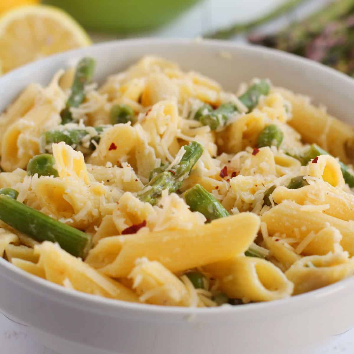 A bowl of Lemon Asparagus Pasta features penne mixed with asparagus and topped with grated cheese, with fresh lemon and asparagus visible in the background.