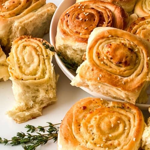 A plate of golden-brown garlic rolls, some topped with sesame seeds, arranged with a few sprigs of fresh herbs on a white surface, evokes the tempting look of Pull Apart Pizza Bread.